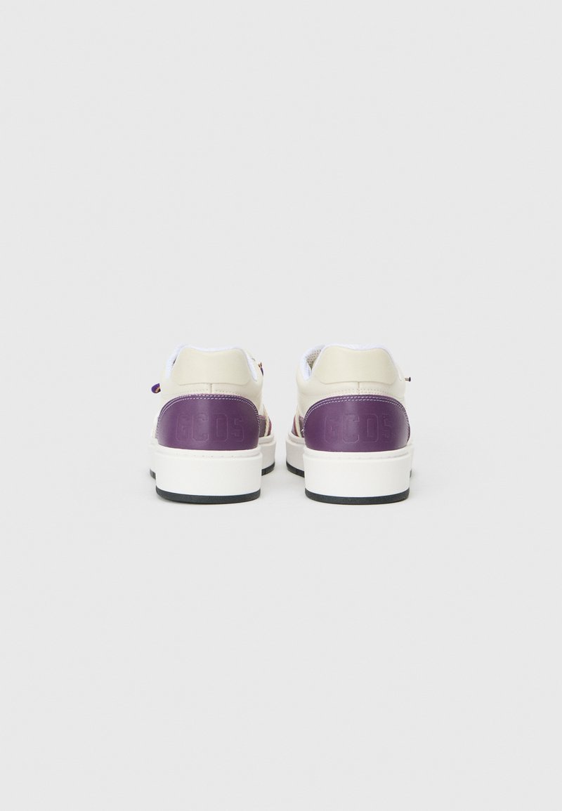Baskets blanches avec un talon et une marque violets brillants, présentant un dessus texturé, une pointe ronde et une semelle blanche épaisse. Lacets circulaires visibles.
