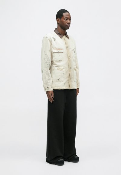 Veste beige clair avec un col marron, dotée de deux poches poitrine et d'une texture usée. Pantalons larges noirs.