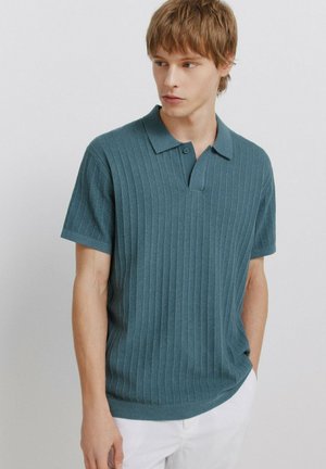 Ung mand med lyst brunt hår iført en grønblå ribstrikket kortærmet polo og hvide bukser, stående mod en ensartet lys baggrund.