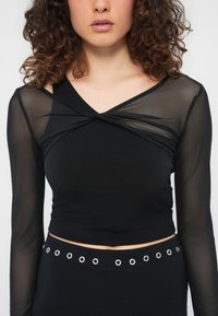 Γυναίκα με σγουρά μαλλιά που φοράει μαύρο διαφανές crop top με μακριά μανίκια και στριμμένο στο στήθος, καθώς και μαύρο παντελόνι με ασημένιους κρίκους στη μέση.