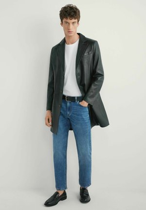 Młody mężczyzna w czarnym skórzanym płaszczu, białej koszulce, niebieskich dżinsach, czarnym pasku i czarnych mokasynach stojący na tle jasnego, jednolitego tła.