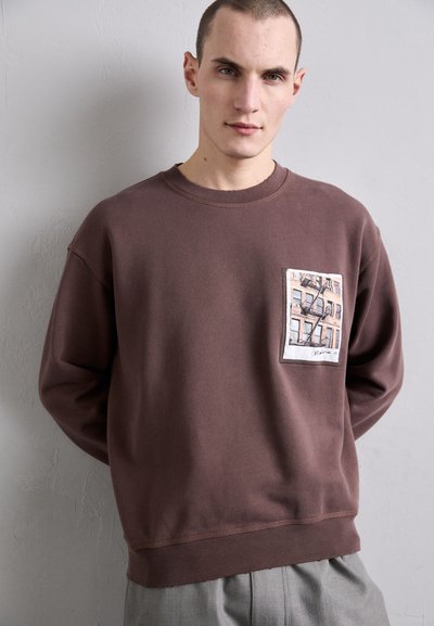 Sweatshirt castanho feito de algodão com um corte descontraído. Apresenta uma impressão de um edifício na bolsa frontal e punhos e barra canelados.
