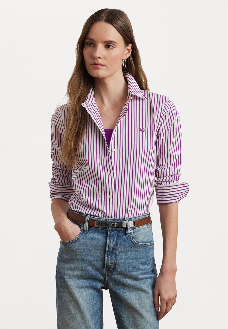 Camicia a righe viola e bianche con una consistenza morbida, maniche arrotolate e un logo visibile, abbinata a jeans in denim azzurro chiaro e una cintura marrone.