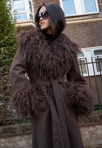 Rudas paltas su pūkine, kailinę primenančia apykakle ir rankovių akcentais. Audinys atrodo minkštas, su surištu liemeniu ir sagų detalėmis.