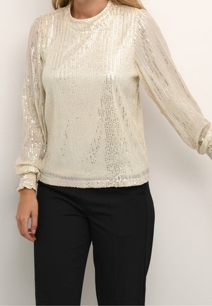 Femme portant un haut crème à manches longues avec sequins, poignets froncés et pantalon noir, debout devant un fond clair uni.