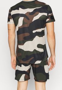 Zestaw odzieży sportowej w kamuflażowym wzorze w stonowanych odcieniach zieleni, brązu i beżu. Bluzka ma krótkie rękawy; szorty sięgają do kolan.