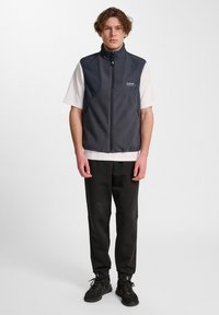 Junger Mann steht da und trägt eine dunkle ärmellose Fleeceweste über einem weißen kurzärmeligen Hemd, schwarze Hosen und schwarze Sneakers vor einfarbigem Hintergrund.