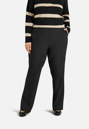Personne portant un pantalon noir à jambes larges et un haut rayé noir et beige à manches longues, les mains dans les poches, debout sur fond blanc.