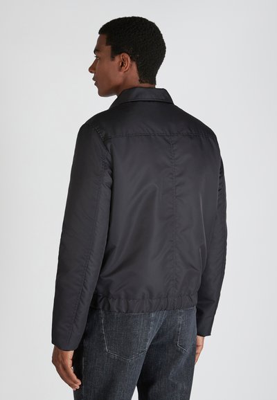 Giacca leggera nera con una texture liscia, colletto e orlo elastico, vista da dietro, abbinata a pantaloni in denim scuro.