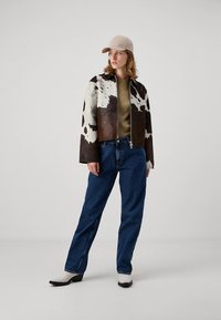 Ruskea-valkoinen lehmännahkatakki, vetoketjullinen edestä, päällä oliivinvihreä paita, siniset farkut ja beige lippalakki, viimeistelty mustavalkoisilla kengillä.