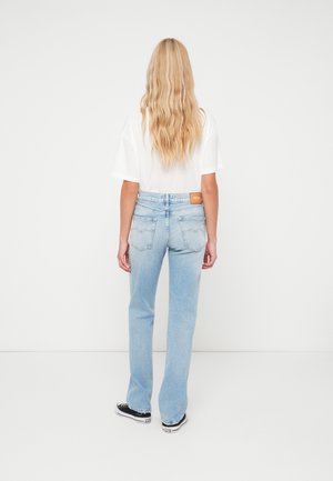 Ανοιχτό μπλε ψηλόμεσα τζιν με ίσια γραμμή, μια ετικέτα με το λογότυπο στο πίσω μέρος και λεία υφή τζιν. Φορεμένα με ένα λευκό μπλουζάκι.