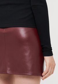 Burgundy faux leather mini skirt with a glossy finish, featuring a fitted design and a seam detail. Paired with a black ribbed long-sleeve top.