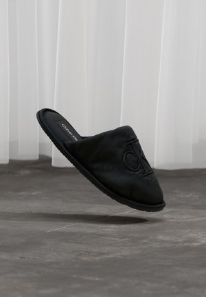 Scarpa nera slip-on con tomaia in tessuto morbido e suola piatta in gomma. Presenta un logo prominente sulla parte anteriore e una finitura texturizzata.