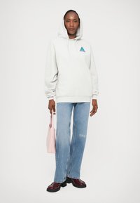 Grijze hoodie met een driehoekig blauw logo, gecombineerd met lichtblauwe jeans met witte tekst. Accessoires zijn een roze tas en bordeauxrode schoenen.