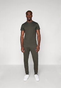 Tumši zaļš kokvilnas T-krekls un saskaņoti sporta bikses ar piegulošu dizainu, kas raksturotas ar apaļu kakla izgriezumu un baltām sporta apaviem. Vienkāršs, saskaņots izskats.