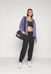 Lila sportjacka, svart kort topp, svarta mjukisbyxor med vita ränder, vita skor och en svart Adidas-duffelväska.