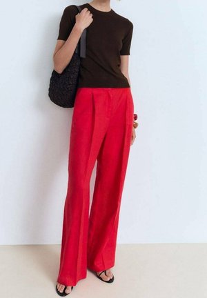 Personne portant un haut noir à manches courtes, un pantalon rouge taille haute à jambes larges, des sandales noires et portant un sac à bandoulière noir texturé.