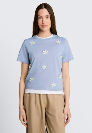 Jonge vrouw met steil bruin haar, gekleed in een lichtblauw shirt met korte mouwen met witte bloemapplicaties en beige broek.