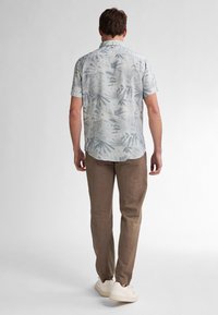Camicia a maniche corte di colore azzurro chiaro con stampa di foglie di palma. Abbinata a pantaloni marroni chiari e sneaker bianche, vista da dietro.