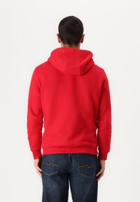 Sudadera roja con capucha hecha de material suave, con dobladillo y puños acanalados, combinada con jeans azul oscuro, vista desde atrás.