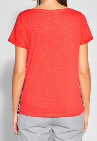 Červený krátký rukáv s volným střihem a kulatým výstřihem, s pruhovanými bočními panely pro kontrast s hladkou texturou.