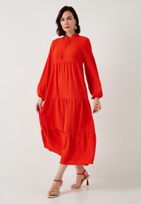 Punainen kerroksellinen maxi-mekko, jossa on pitkät hihat, nauhakatkaisu ja tekstuuroitu kangas. Yhdistetty monivärisiin remmisandaaleihin.