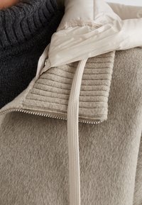 Helle beigefarbene Fleecejacke aus strukturiertem Stoff, mit cremefarbener Steppkapuze und gerippten Akzenten. Sie verfügt über einen Reißverschluss und eine weiche Oberfläche.