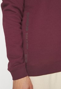 Bordowa bluza z prążkowanymi mankietami i dołem, z delikatnym szarym napisem marki wzdłuż rękawa. Wykonana z miękkiej, teksturowanej tkaniny.