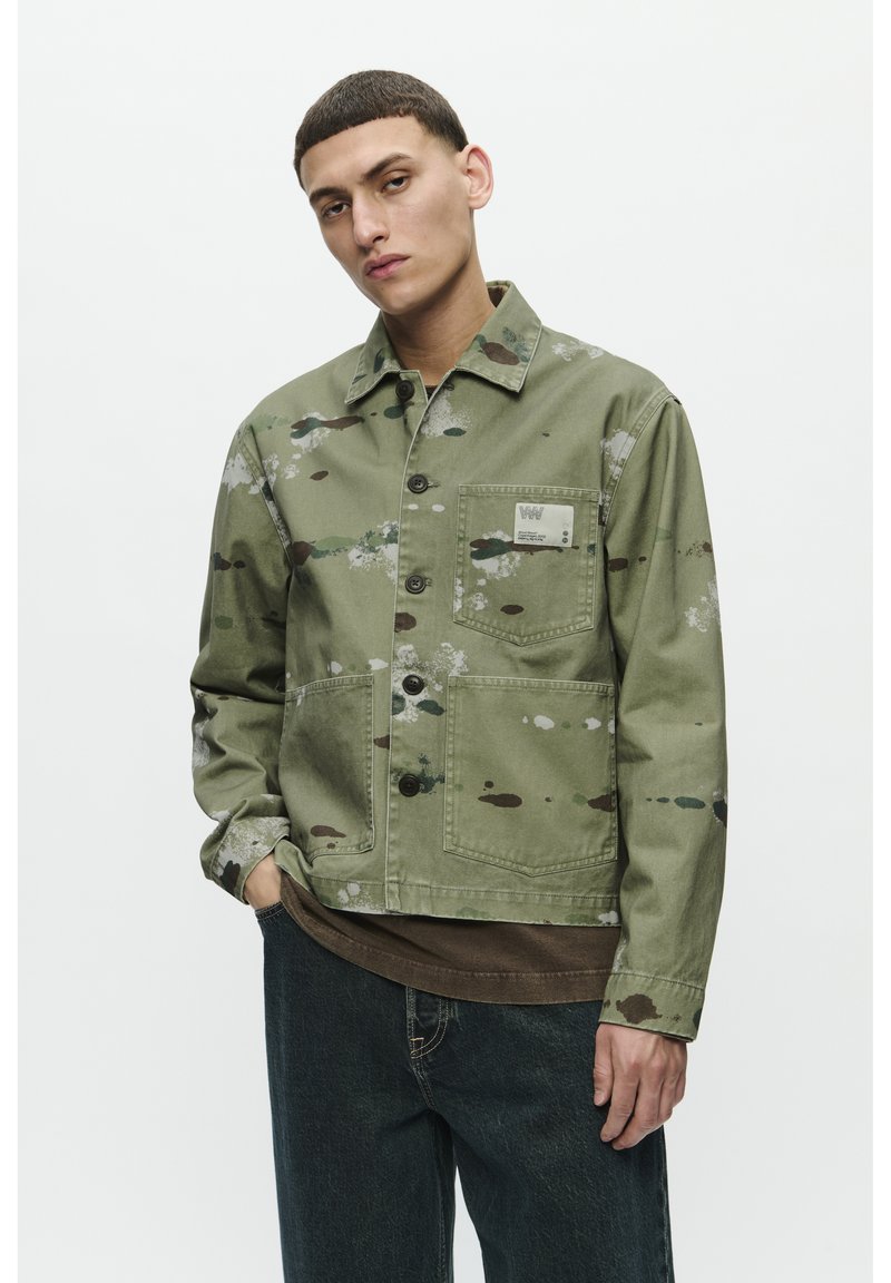 Chaqueta de lona verde con un patrón salpicado, cierre de botones, bolsillo en el pecho y cuello. Combinada con una camisa marrón y jeans azul oscuro.