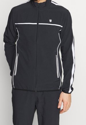 Mann trägt eine schwarze Trainingsjacke mit Reißverschluss und weißen Streifen an den Ärmeln, der Brust und den Taschen, kombiniert mit passenden schwarzen Hosen.