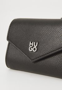 Bolso de mano de cuero negro texturizado con cierre de solapa. Presenta un logo plateado "HUGO" en la parte frontal. Acabado suave con un diseño estructurado.