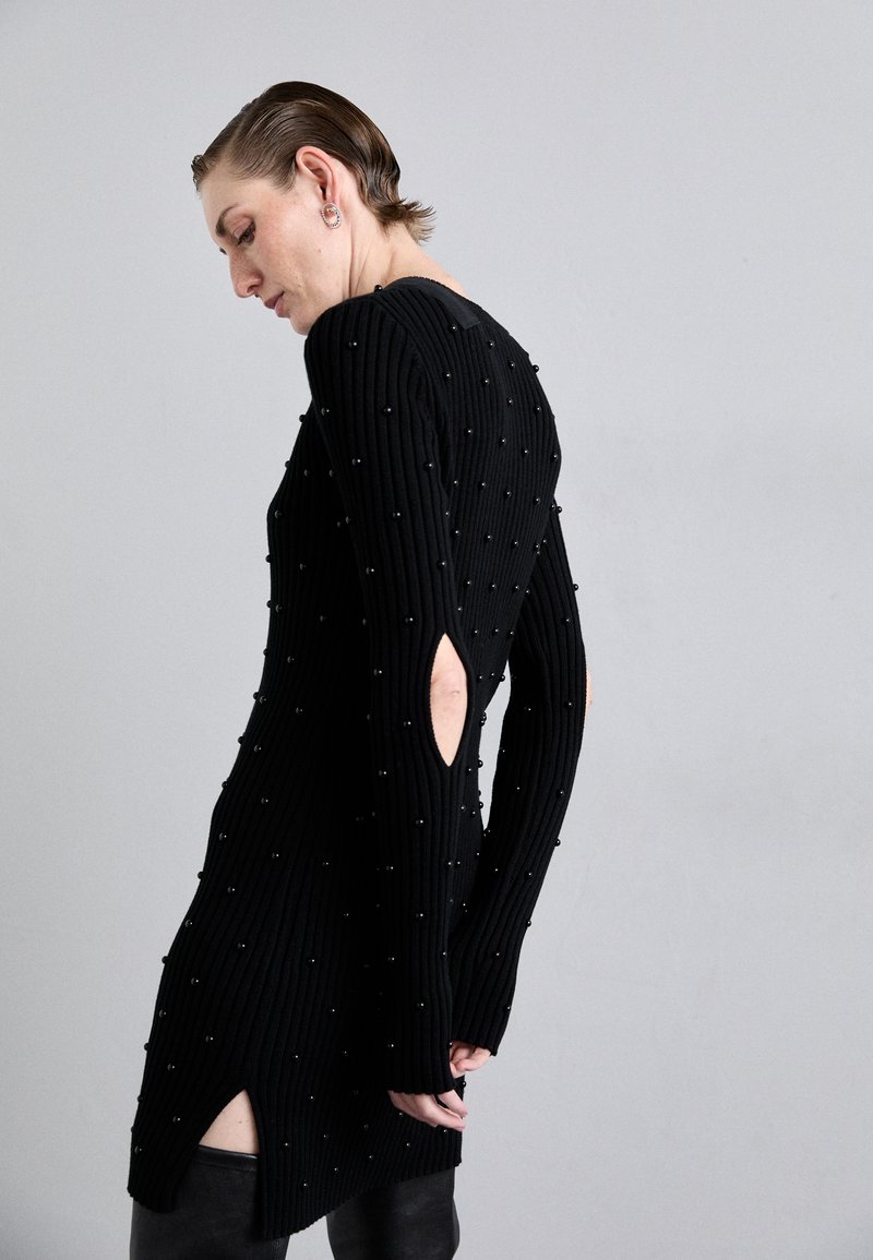 Robe noire à côtes ornée de petites perles, avec des manches longues, des découpes sur les côtés et un col haut. Fabriquée en tissu tricoté.