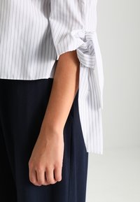 Chemise rayée blanche et marine avec un détail de cravate sur la manche, mettant en valeur un tissu lisse et une coupe décontractée. Main reposant sur un matériau marine.