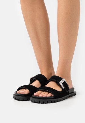 Feet wearing black suede sandals with thick soles and large silver buckles on white background.
