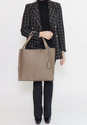 Große beige Leder-Shopper-Tasche mit zwei Griffen, die eine strukturierte Form und einen kleinen angebrachten Anhänger aufweist. Kombiniert mit einem schwarzen Outfit und einem karierten Blazer.