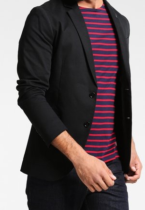 Hombre con blazer negro sobre camisa de rayas rojas y azul marino, con pantalones oscuros, frente a un fondo blanco liso.