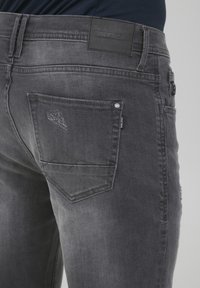 Jeans de mezclilla gris con un acabado texturizado, con un bolsillo trasero que presenta un detalle desgastado sutil y una etiqueta de cuero negro de la marca en la cintura.