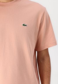 Tricou din bumbac roz deschis, cu mâneci scurte și guler rotund. Prezintă un mic logo brodat cu crocodil verde și alb pe piept.
