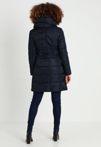 Mörkblå pufferjacka med hög krage, insnörning i midjan och kviltad textur, som sträcker sig under knäna och har långa ärmar.