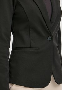 Černý blazer s elegantním designem, texturovanou látkou, zapínáním na jeden knoflík a boční kapsou s diskrétním ozdobným detailem.