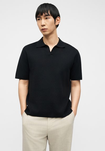 Νέος άνδρας με μαύρο κοντομάνικο πόλο πουκάμισο και ανοιχτό μπεζ παντελόνι, στέκεται με τα χέρια στις τσέπες, κοιτάζοντας ελαφρώς προς τα αριστερά σε απλό φόντο.