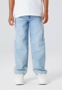 Bambino vestito con jeans azzurri larghi, sneakers bianche e una maglietta bianca, in piedi contro uno sfondo chiaro e uniforme.