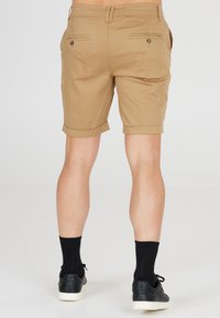 Khaki shorts met een iets taps toelopende pasvorm, opgerolde manchetten, twee achterzakken en zichtbare stiksels. Draagt met zwarte sokken en sneakers.