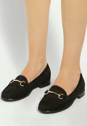 Black suede loafers with a rounded toe, featuring a gold metal accent across the front and a low stacked heel. Soft texture.