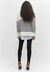 Personne aux longs cheveux bouclés vue de dos, portant un pull crème à rayures noires, des leggings noirs et des talons hauts noirs à brides.