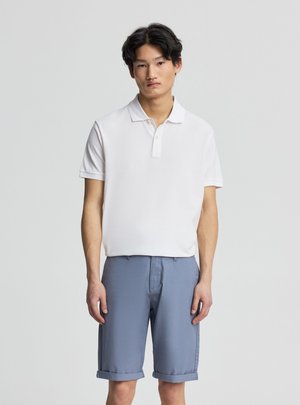 Giovane uomo con capelli neri che indossa una polo bianca a maniche corte e pantaloncini blu risvoltati, in piedi davanti a uno sfondo chiaro e uniforme.