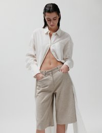 Γυναίκα με βρεγμένα μαλλιά που φοράει ανοιχτό λευκό πουκάμισο με μακριά μανίκια και μπεζ ριγέ σορτς μέχρι το γόνατο, με τα χέρια στις τσέπες, κοιτάζοντας κάτω.