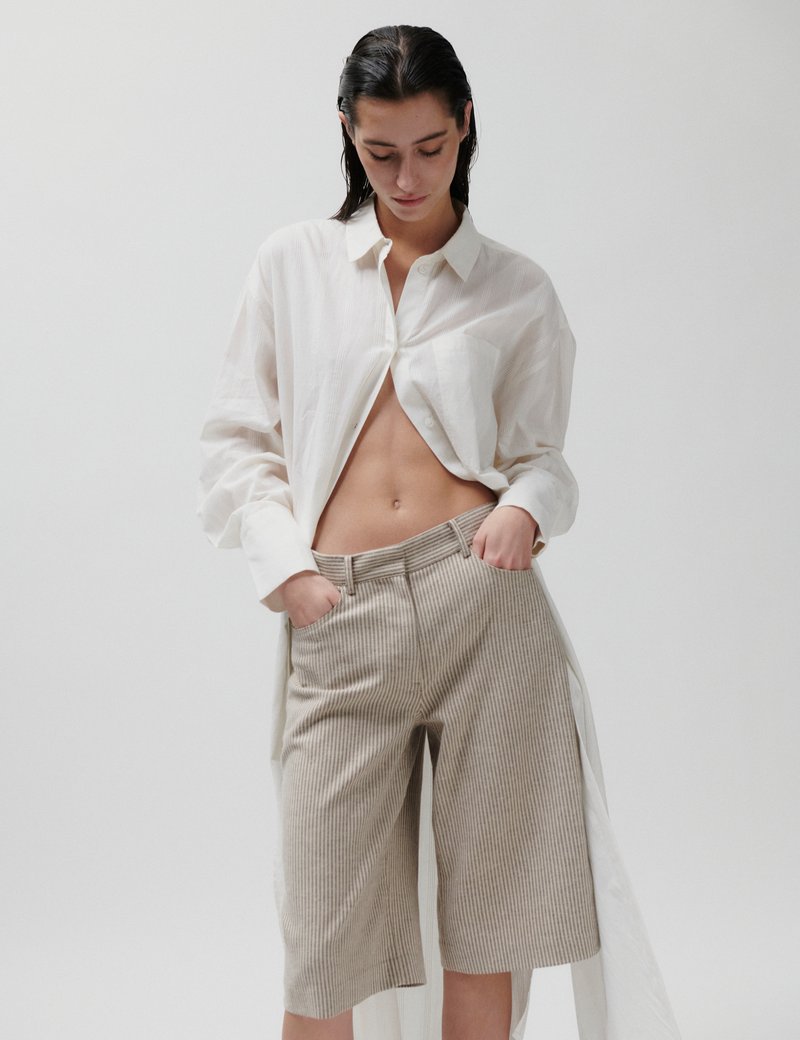 Γυναίκα με βρεγμένα μαλλιά που φοράει ανοιχτό λευκό πουκάμισο με μακριά μανίκια και μπεζ ριγέ σορ τς μέχρι το γόνατο, με τα χέρια στις τσέπες, κοιτάζοντας κάτω.