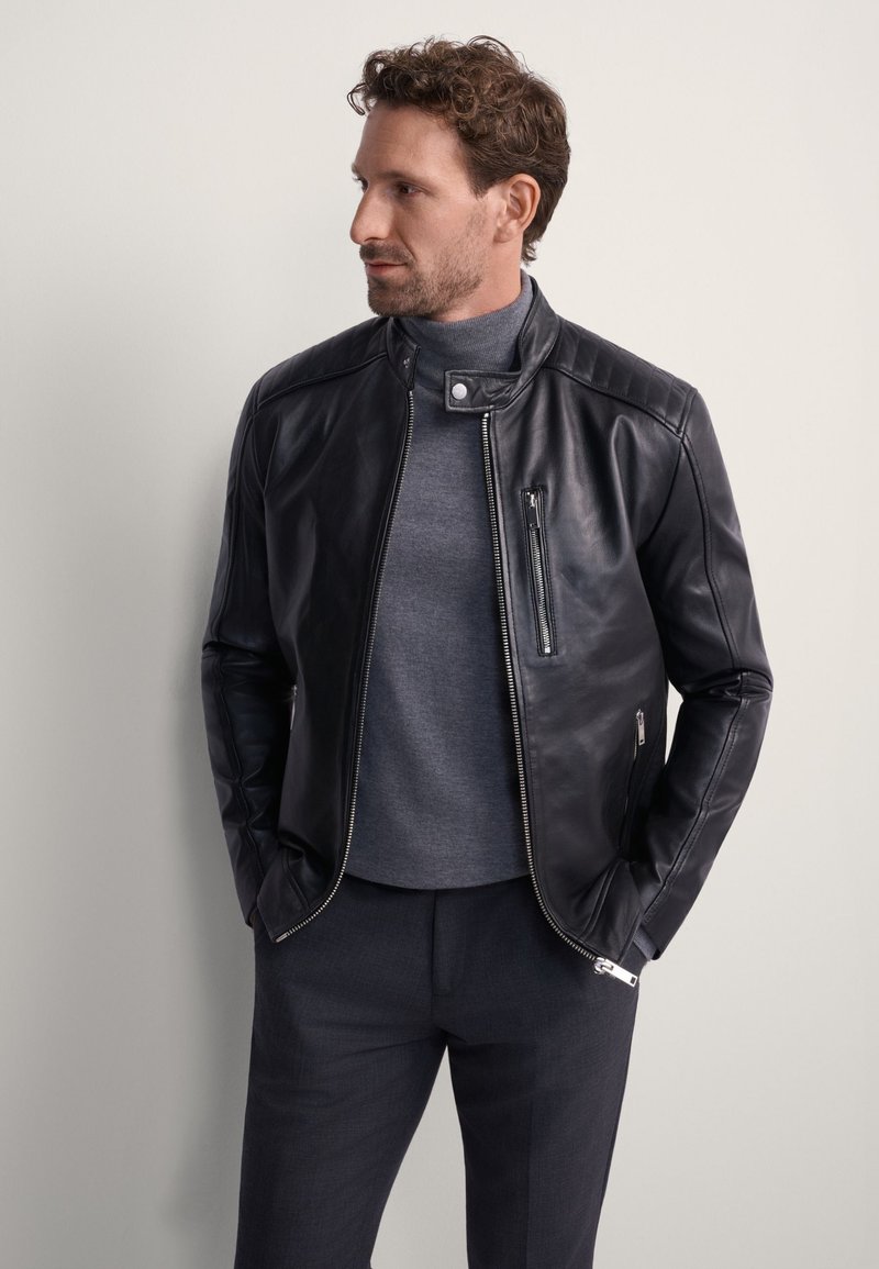 Czarna skórzana kurtka z pikowanym wzorem na ramionach, z zamkiem błyskawicznym z przodu i kieszeniami po bokach. Nosi się ją na szarym golfie i ciemnych spodniach.