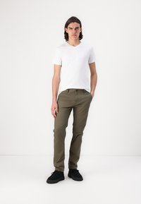 Valkoinen V-kauluksinen t-paita, oliivinvihreät slim-fit-housut, mustat kengät; seisova malli yksinkertaisella taustalla, kädet taskuissa, neutraali ilme.